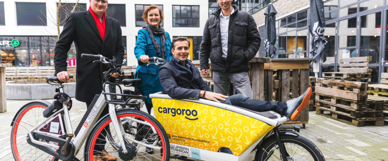 Groepsportret van vier mensen van projectontwikkelaar AM in Delft die poseren met een eletrische fiets en een bakfiets voor vrachtvervoer, waar één van de vier geportretteerden is gaan zitten