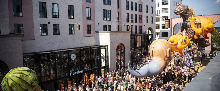 Foto van de feestelijke opening van NS Station Heerlen. We zien een drukke vrolijke optocht voor het station met grote poppen van draken en dinosaurussen