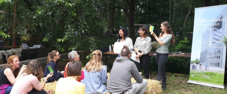 In een groene tuin zien we publiek zitten op hooibalen terwijl drie dames van Wageningen University stellingen voorlezen waar het publiek op reageert door gekleurde kaarten omhoog te houden