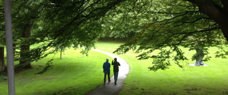 We zien twee mensen wandelend en pratend op een voetpad door een bos. Het beeld is ingekaderd door een grote boom die over het pad en de wandelaars buigt